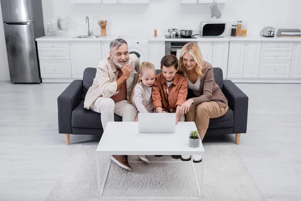 Kids using laptop near happy grandparents on couch in kitchen — Stock Photo