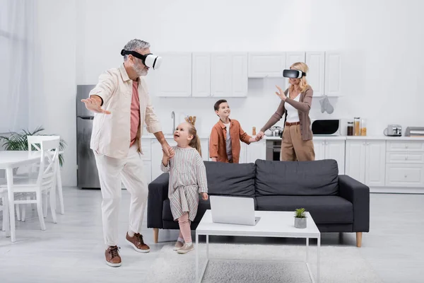 Pareja en auriculares vr tomados de la mano con nietos mientras juega en la cocina - foto de stock