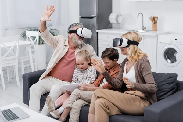 Весёлая пара жестикулирует в наушниках vr рядом с внуками, сидящими на диване с цифровым планшетом — стоковое фото