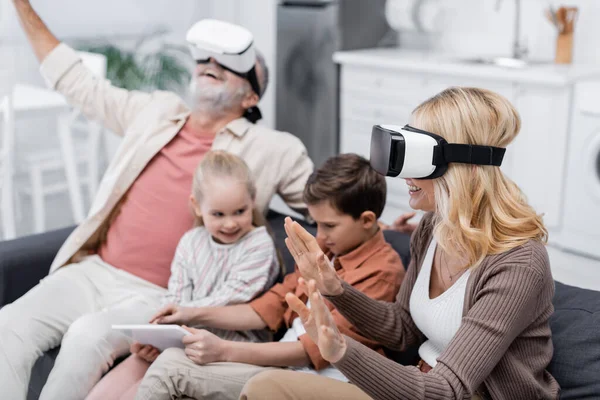 Excited couple gaming in vr headsets near grandchildren using digital tablet on couch — Stock Photo