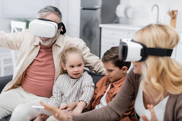 Kinder sitzen auf Sofa mit digitalem Tablet in der Nähe der Großeltern und spielen in vr-Headsets — Stockfoto