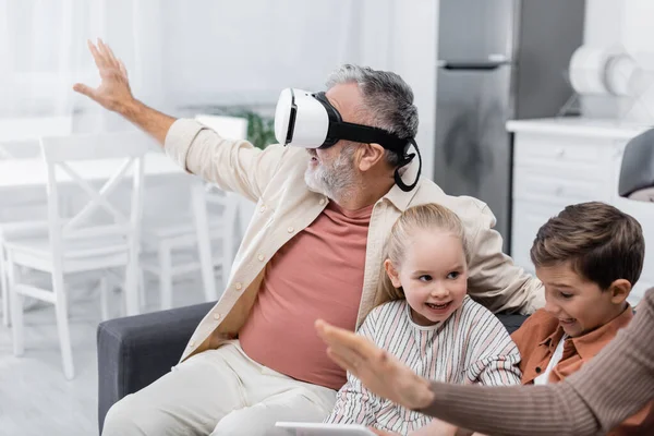 Hombre de edad avanzada en auriculares vr gesto cerca de nietos con tableta digital - foto de stock