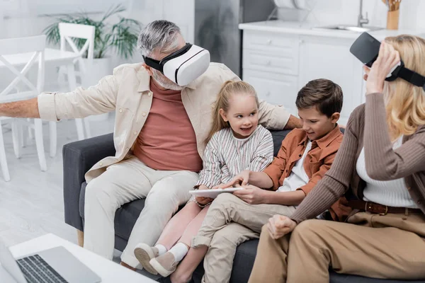 Boy pointing at digital tablet while sitting on couch near sister and grandparents in vr headsets — Stock Photo