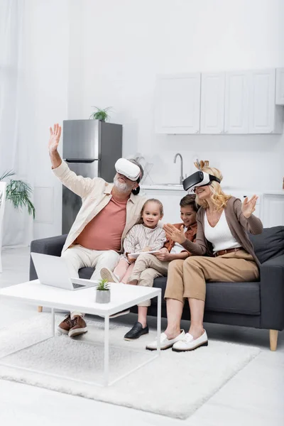 Aufgeregtes Paar gestikuliert beim Spielen in vr Headsets in der Nähe von Enkeln mit digitalem Tablet auf dem Sofa — Stockfoto