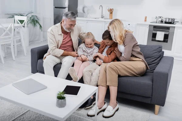 Couple s'amuser avec les petits-enfants assis sur le canapé près de l'ordinateur portable et tablette numérique sur la table — Photo de stock