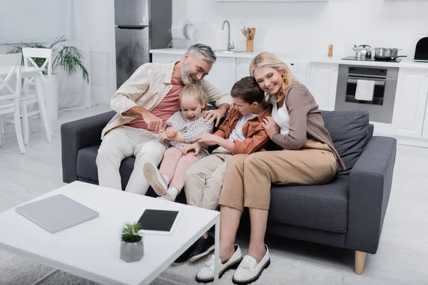 Alegre pareja abrazando nietos cerca de portátil y tableta digital con pantalla en blanco en la mesa - foto de stock