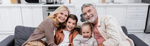 Crianças sorridentes com avós olhando para a câmera no sofá em casa, banner — Fotografia de Stock