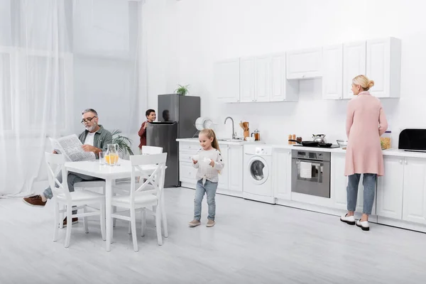 Hombre mayor leyendo periódico cerca de nietos y esposa en la cocina moderna - foto de stock