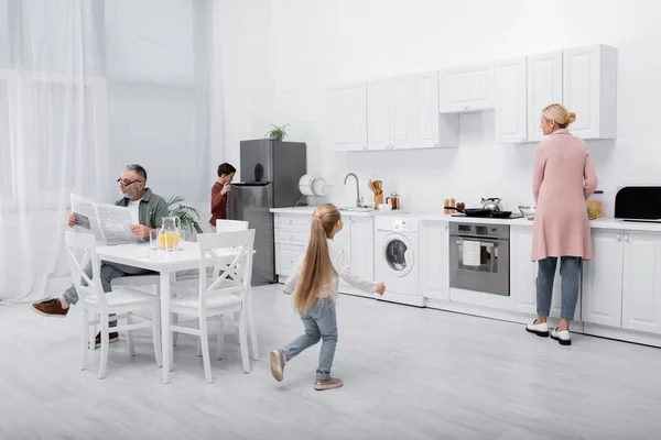 Senior man reading newspaper while grandchildren helping granny preparing breakfast — Stock Photo