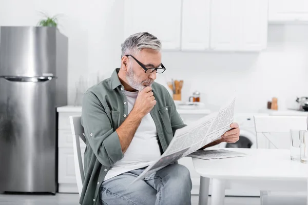 Thoughtful senior man in eyeglasses reading morning newspaper in kitchen — Stock Photo