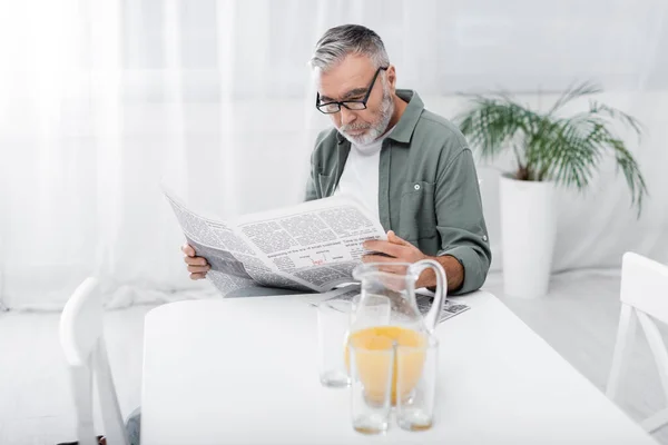 Старший мужчина в очках читает утреннюю газету возле кувшина с апельсиновым соком — стоковое фото