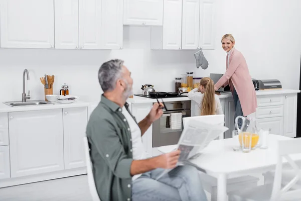 Senior homme avec journal et lunettes regardant femme heureuse préparer le petit déjeuner avec petite-fille — Photo de stock