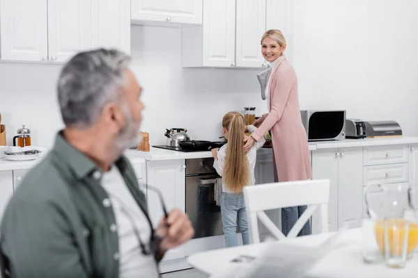 Femme souriante regardant mari flou tout en préparant le petit déjeuner avec petite-fille — Photo de stock