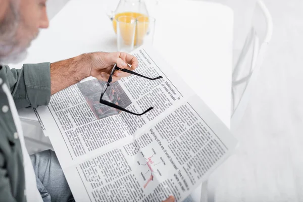 Vista recortada de hombre mayor borrosa sosteniendo gafas mientras lee el periódico - foto de stock