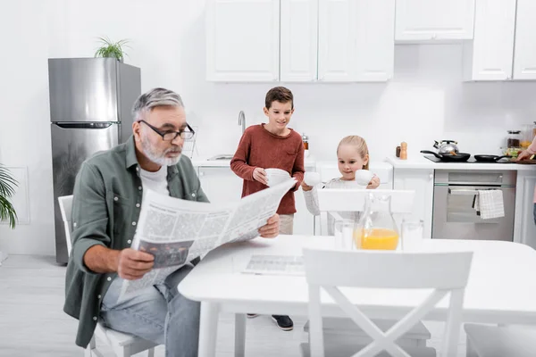 Anziano uomo leggendo giornale mentre nipotini apparecchiare la tavola per la colazione — Foto stock