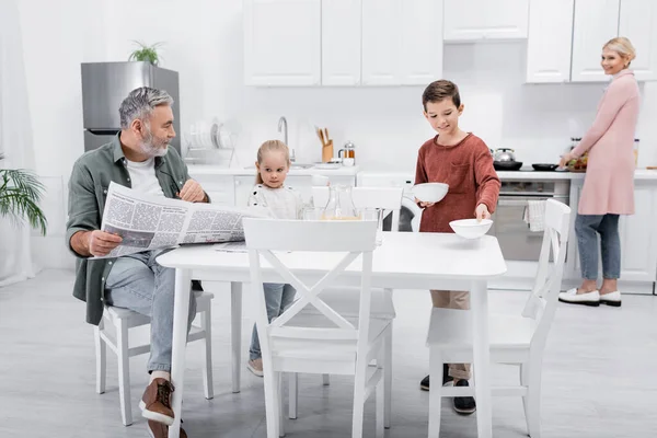 Uomo anziano con giornale guardando nipote che tiene tazze in cucina — Foto stock