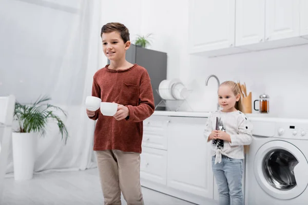 Bambini sorridenti che tengono tazze e posate in cucina — Foto stock