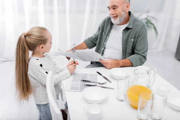 Uomo anziano con giornale che parla con nipote che tiene la forchetta in cucina — Foto stock