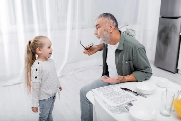 Gray-haired man holding eyeglasses and pointing with hand while talking to granddaughter — Stock Photo