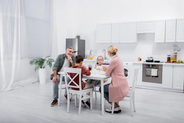 Hombre mayor sonriendo cerca de nietos y esposa desayunando en la cocina moderna - foto de stock