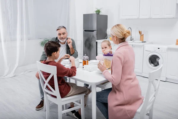 Heureux senior homme regardant petits-enfants et femme prendre le petit déjeuner dans la cuisine — Photo de stock