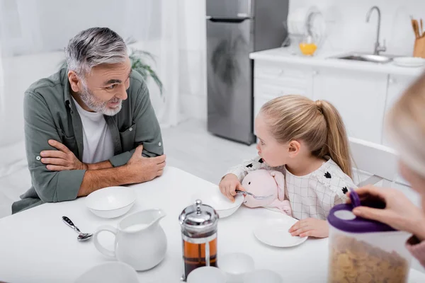 Positive senior man talking to granddaughter near blurred wife with corn flakes — Stock Photo