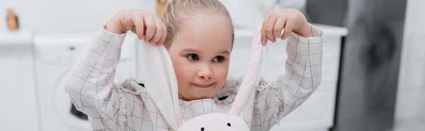 Маленькая девочка играет с игрушечным кроликом на кухне, баннер — стоковое фото