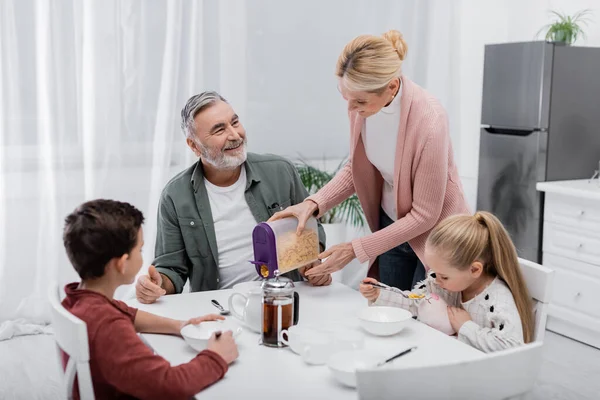 Glücklicher Senior schaut Frau beim Frühstück mit Enkeln beim Servieren von Cornflakes zu — Stockfoto