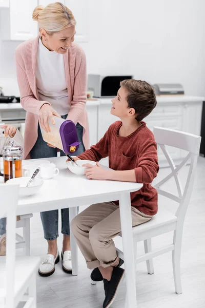 Femme heureuse tenant le récipient avec des flocons de maïs près du petit-fils pendant le petit déjeuner — Photo de stock