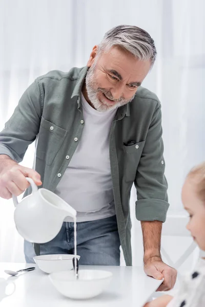 Happy granddad pouring fresh milk near blurred girl in kitchen — Stock Photo