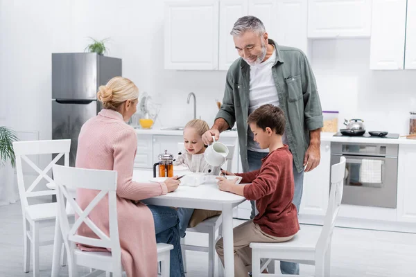 Homme heureux versant du lait pour femme et petits-enfants pendant le petit déjeuner — Photo de stock