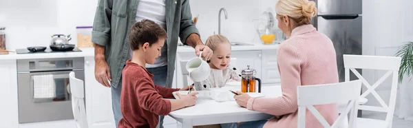 Senior Mann gießt Milch in der Nähe von Enkeln und Frau beim Frühstück in der Küche, Banner — Stockfoto