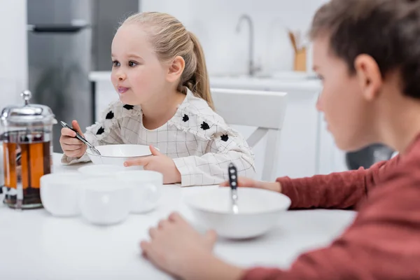 Girl having breakfast near blurred brother in kitchen — Stock Photo