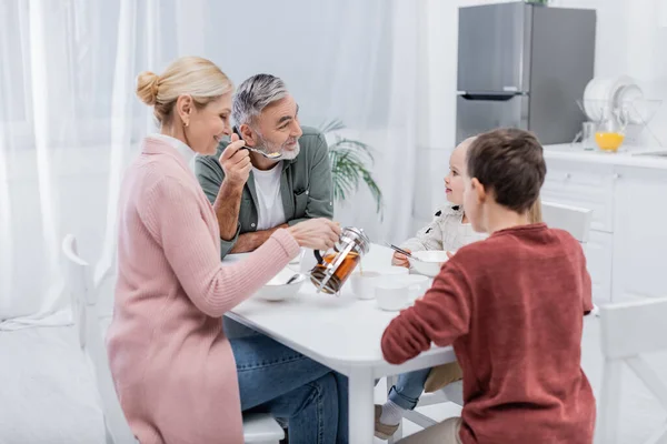 Lächelnde Frau mittleren Alters gießt beim Frühstück Tee in die Nähe von Enkeln und Ehemann — Stockfoto