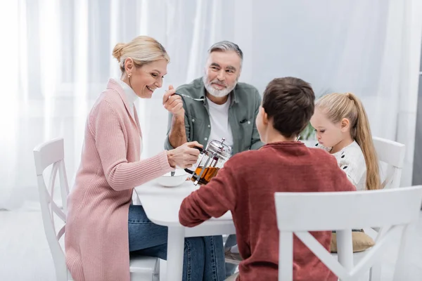 Lächelnde Frau gießt beim Frühstück Milch in die Nähe von glücklichem Ehemann und Enkeln — Stockfoto