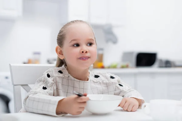 Маленькая девочка ест завтрак, сидя в размытой кухне — стоковое фото