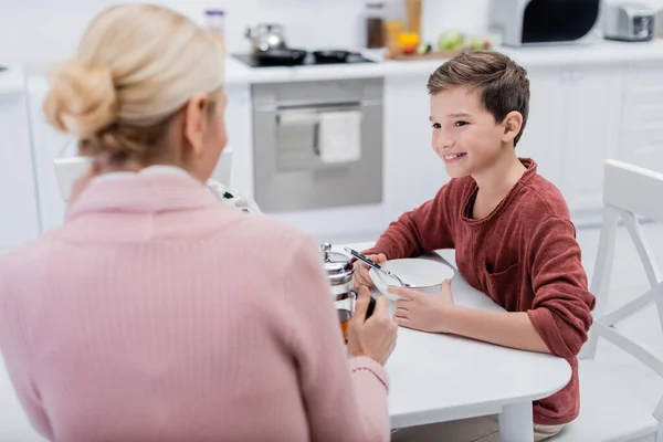 Sorridente ragazzo che fa colazione vicino alla nonna offuscata in cucina — Foto stock