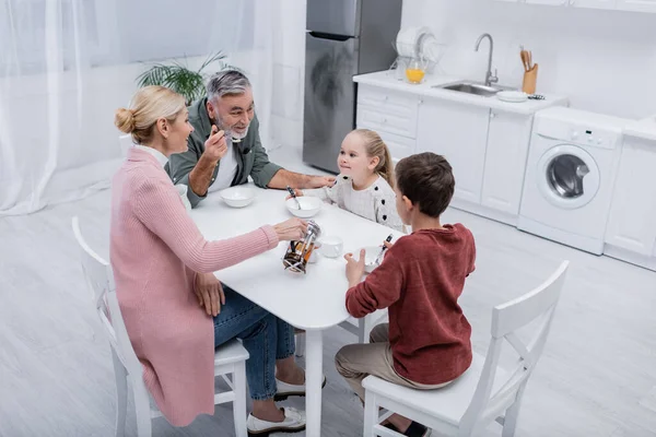 Cheerful man talking to grandchildren near wife pouring tea during breakfast — Stock Photo