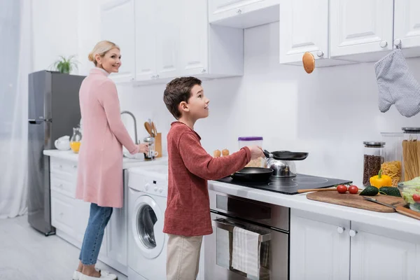 Menino cozinhar panqueca perto de legumes frescos e vovó sorridente na cozinha — Stock Photo