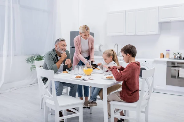 Happy woman serving pancakes for grandchildren and husband during breakfast — Stock Photo
