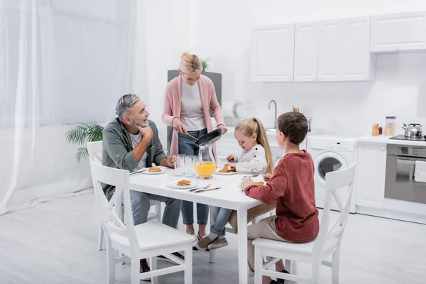 Donna di mezza età con padella vicino marito e nipoti che fanno colazione in cucina — Foto stock