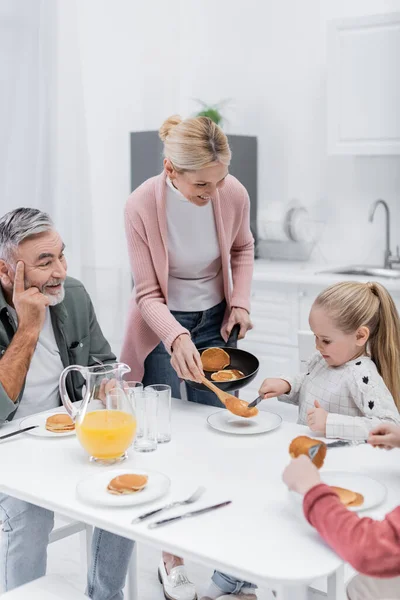 Lächelnde Frau mit Pfanne und Pfannkuchen in der Nähe von Mann und Enkeln — Stockfoto