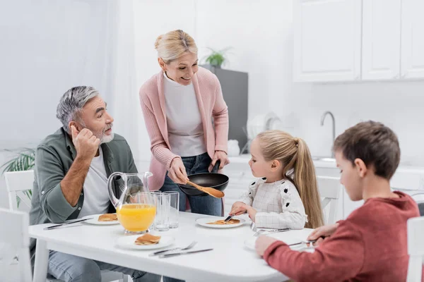 Glückliche Frau hält Pfanne und Spachtel in der Nähe von Enkeln und Ehemann essen Pfannkuchen zum Frühstück — Stockfoto