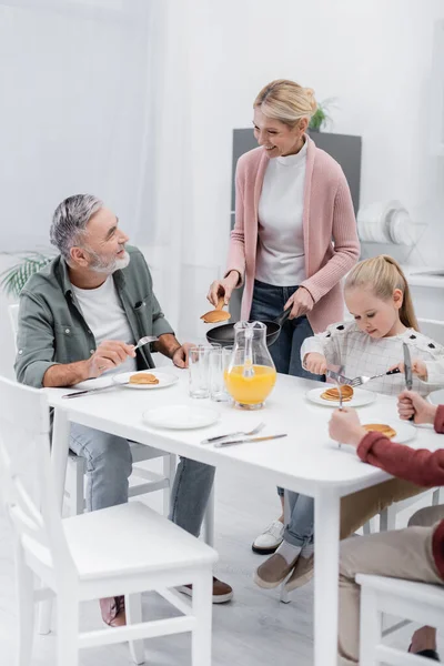 Mujer de mediana edad sosteniendo sartén con panqueques cerca de marido feliz y nietos - foto de stock