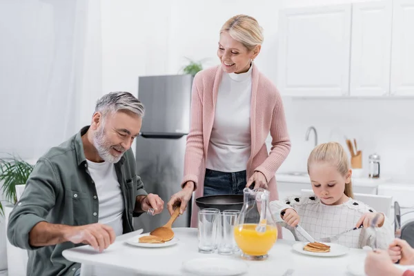 Happy woman serving pancakes for husband during breakfast with grandchildren — Stock Photo