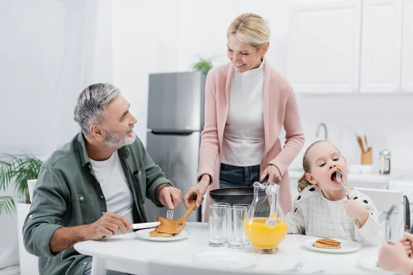 Cheerful woman serving pancakes for husband during breakfast with grandchildren — Stock Photo