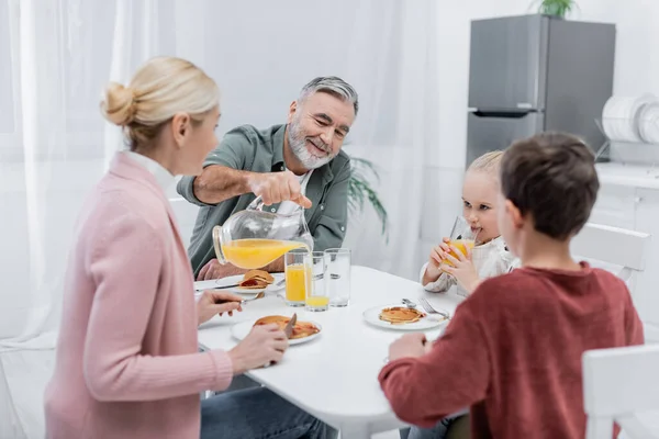 Hombre mayor que vierte jugo de naranja durante el desayuno con esposa y nietos - foto de stock
