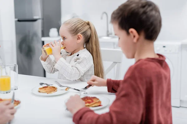 Girl drinking orange juice near pancakes and blurred brother — Stock Photo