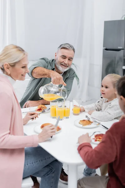 Sorridente homem sênior derramando suco de laranja para esposa e netos durante o café da manhã — Fotografia de Stock