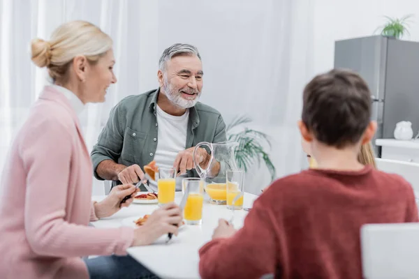 Homem sênior alegre tomando café da manhã com netos desfocados e esposa na cozinha — Fotografia de Stock